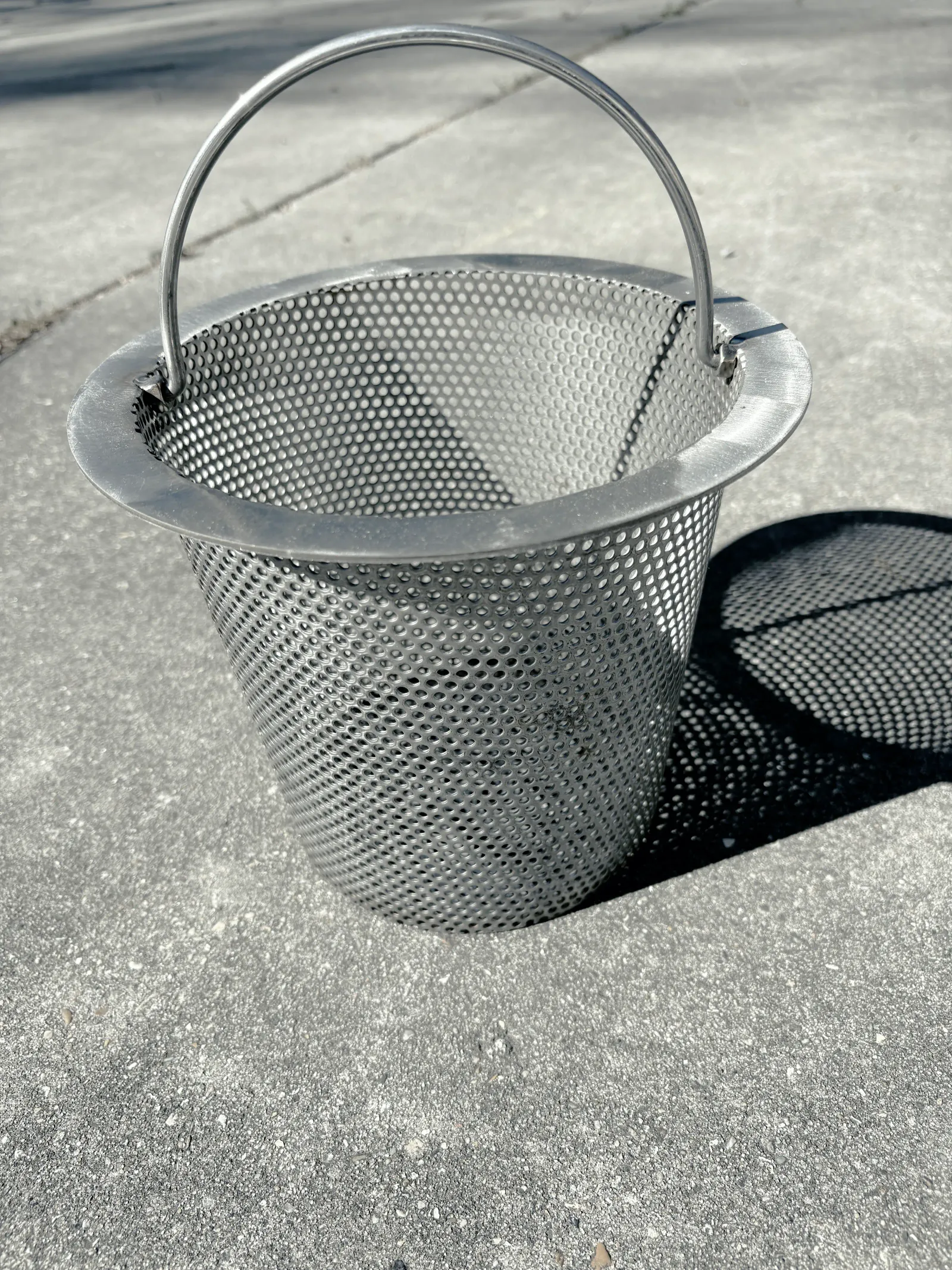 Whole stainless steel skimmer basket, handle up, on sunlit concrete, its dotted shadow beside it.