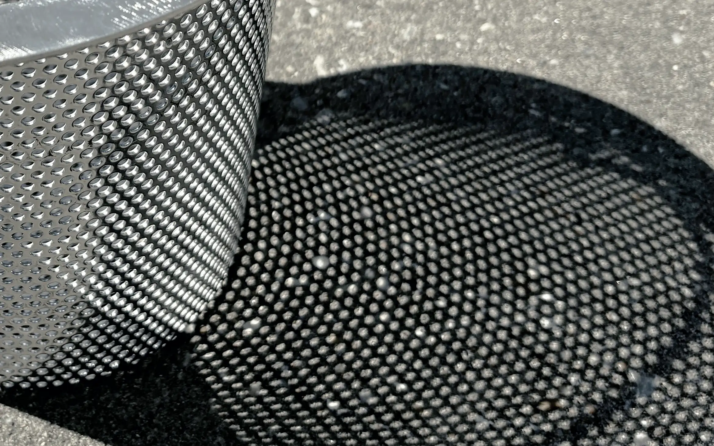 The dotted shadow of a stainless steel basket's perforated wall, on sunlit concrete.