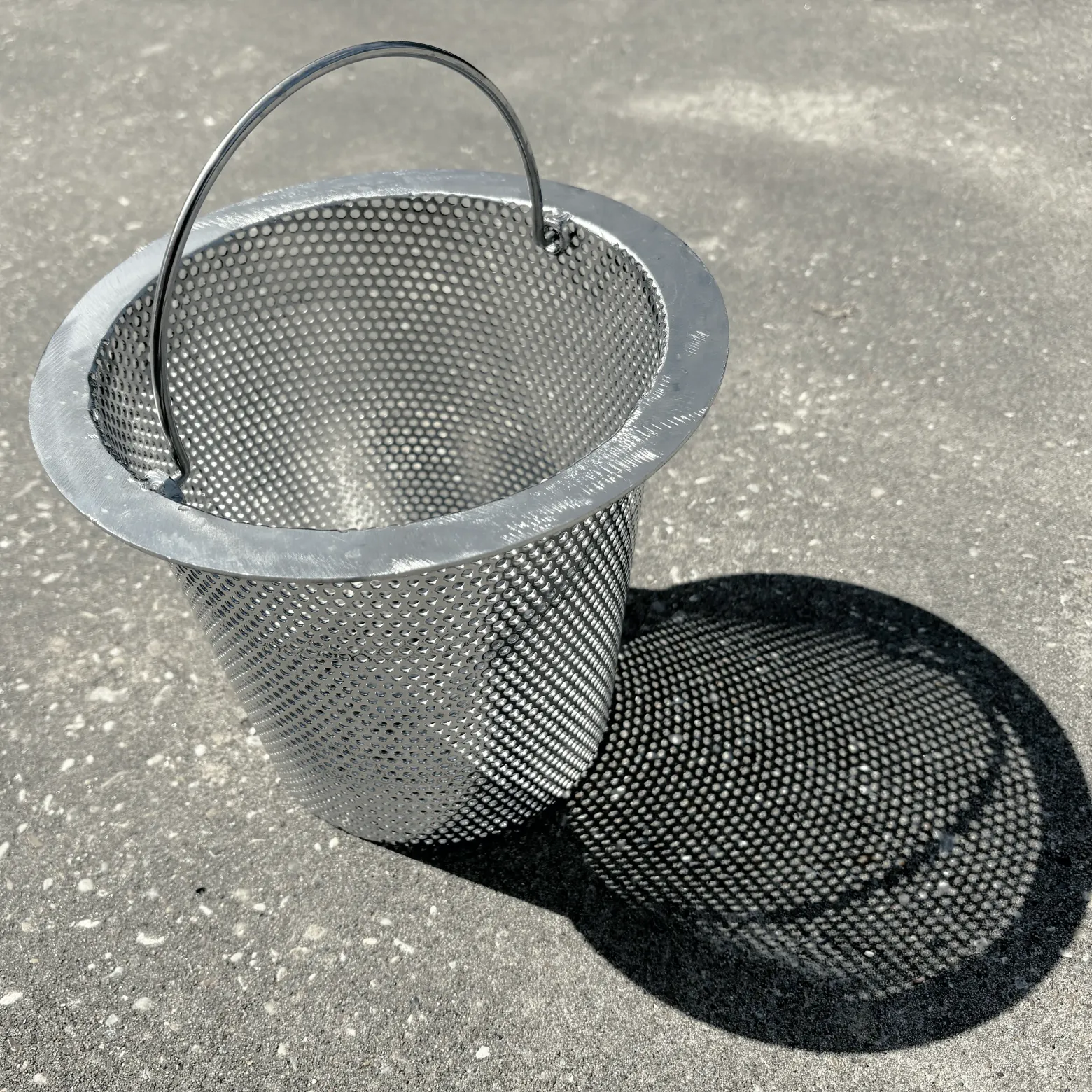 A whole stainless steel skimmer basket, handle up, on sunlit concrete, with its whole dotted shadow beside it.