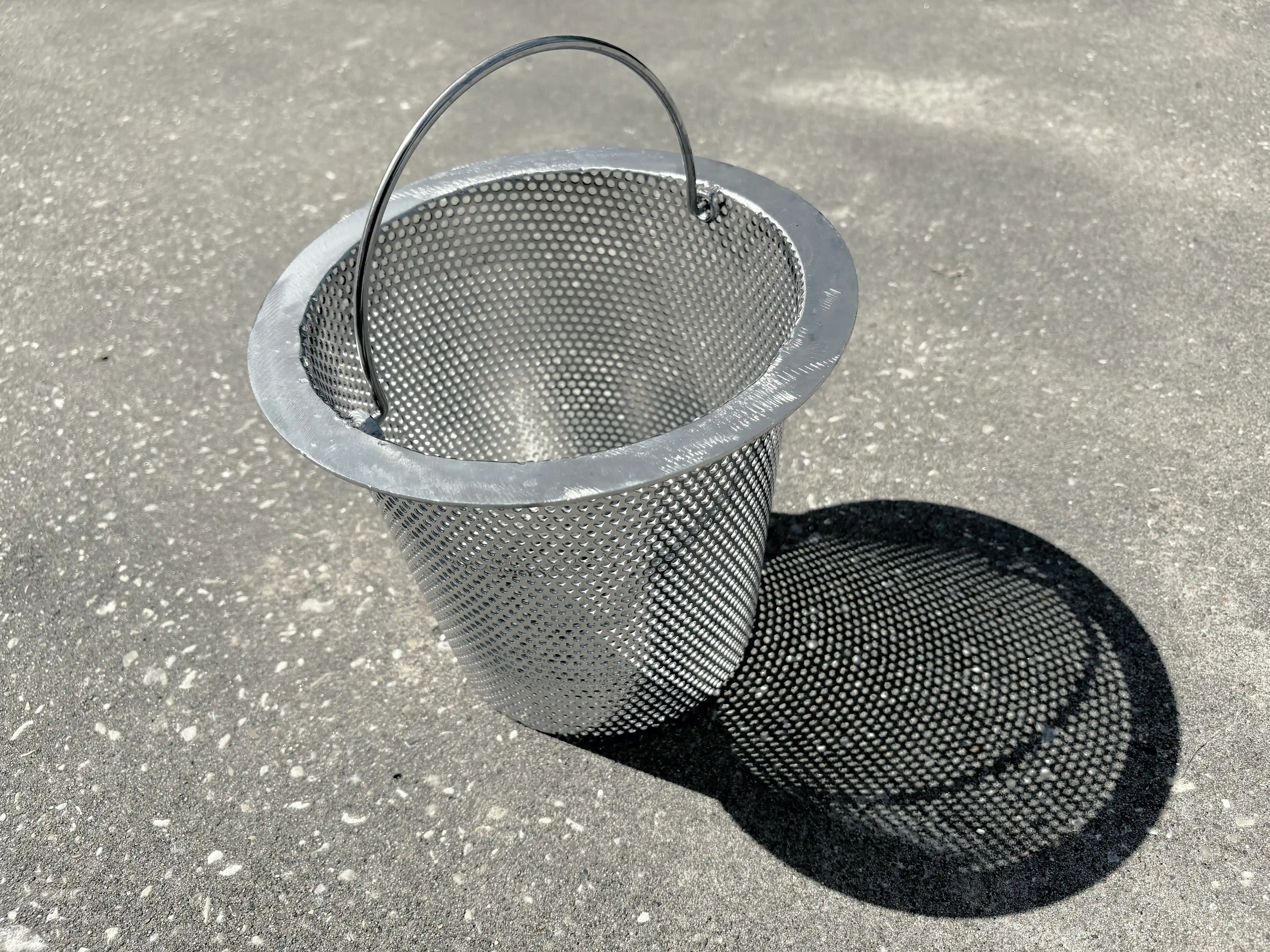 A whole stainless steel skimmer basket, handle up, on sunlit concrete, with its whole dotted shadow beside it.
